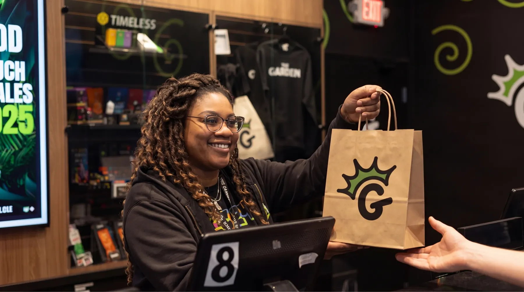 A smiling Garden budtender handing a customer a paper bag printed with the crowned G