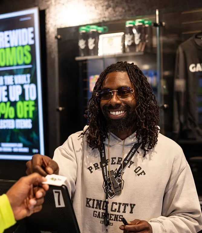 A smiling Garden budtender in a Property of King City Gardens hoodie, taking a customer's card at the checkout counter